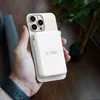 A hand holds a smartphone with a white power bank attached to the back. A plant is visible in the background. Náhled obrázku produktu