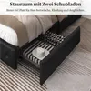A black upholstered bed frame with open storage drawers showing bedding and a hat inside. Produktbild-Vorschau