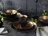 A table setting with black and gold dinnerware, including plates, bowls, and a mug, with some greenery. Náhľad obrázku produktu