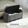 A gray upholstered storage bench with its lid open, placed next to a bed in a bedroom setting. A white fluffy rug is in the foreground. Anteprima dell’immagine prodotto