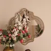 A decorative mirror with a unique shape is shown on a wall, reflecting a lamp and a palm leaf. In the foreground, a vase of flowers, including cotton bolls and pink roses, is arranged. Produktbild-Vorschau