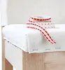 A close-up view of a white fabric seat cushion on a wooden chair, with a roll of red and white patterned ribbon placed on top. Produktbild-Vorschau