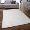 A white shaggy rug is placed on a wooden floor in front of a modern sofa and a cabinet. Produktbild-Vorschau