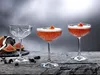 Three coupe glasses on a wooden surface, two filled with a red cocktail and one empty, with grapefruit slices and bar tools in the background. Náhled obrázku produktu