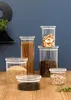 A collection of glass storage jars filled with various dry goods, arranged on a wooden surface against a blue wall. Produktbild-Vorschau