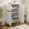 A white shoe cabinet with its doors open, displaying shoes and other items on its shelves. The cabinet is placed on a wooden floor next to a basket and a rug. Produktbild-Vorschau