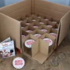 A cardboard box filled with jars, with a booklet and a loose lid on a wooden surface. Produktbild-Vorschau