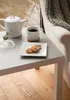 A close-up shot of a white teapot, a white mug of coffee, and two cookies on a white plate, all placed on a light gray table next to a cream-colored armchair with a green knitted blanket draped over it. The background shows a window with a blurred view of  Produktbild-Vorschau