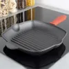 A cast iron grill pan with a red silicone handle protector sits on a black glass stovetop. In the background, jars of food items are visible on a shelf. Voir les images du produit