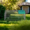 A metal cage with a green roof sits on a grassy lawn in front of a small building. Náhled obrázku produktu