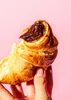 A hand holding a chocolate-filled croissant against a pink background. Produktbild-Vorschau