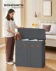A woman is putting clothes into a large, gray, multi-compartment laundry basket in a bedroom. Produktbild-Vorschau