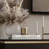 A close-up of decorative items on a black and gold dresser, including a vase with dried flowers, a gold candle holder with white candles, and stacked books. Produktbild-Vorschau