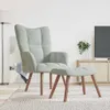 A light gray velvet armchair and matching ottoman are placed on a rug in a living room setting, with a TV stand and a plant in the background. Náhľad obrázku produktu
