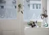 A window decorated with static window film featuring a winter forest scene with deer and trees, along with festive decorations like pine cones, ornaments, and candles on the windowsill. Produktbild-Vorschau
