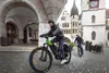 Two people riding electric mountain bikes in a European town square with historic buildings. Produktbild-Vorschau
