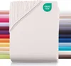 A stack of folded fitted sheets in various colors, with a white fitted sheet prominently displayed in the foreground. A green OEKO-TEX logo is visible on the white sheet. Produktbild-Vorschau
