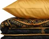 A stack of bedding, including a pillow and a folded duvet or comforter, with a gold pillow on top and a black and gold patterned duvet below. Produktbild-Vorschau