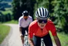 Two cyclists riding on a road with trees and hills in the background. Anteprima dell’immagine prodotto