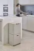 A person is blurred in the background of a modern kitchen, interacting with the sink area. In the foreground, a two-tiered trash and recycling bin is prominently displayed. Podgląd zdjęcia produktu