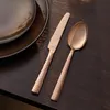 A close-up shot of a rose gold colored knife and spoon set placed on a dark wooden table next to a dark plate. Náhľad obrázku produktu