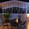 An outdoor patio at night, decorated with white icicle string lights hanging from a canopy. A table with chairs is set up, with bottles and a lantern on the table. Produktbild-Vorschau