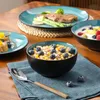 A table setting with various dishes, including a bowl of cereal with blueberries, a plate with steak and asparagus, and a bowl of yogurt with fruit. Voir les images du produit