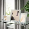 Three glass picture frames with abstract art displayed on a glass table in front of a window with curtains. Produktbild-Vorschau