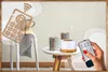 A hand holding a remote control points at an aroma diffuser with candles on a side table, with a graphic of a remote control and Wi-Fi symbol in the background. Produktbild-Vorschau