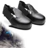 A pair of black safety shoe covers with steel toes, presented on a white background. A faded image of a person using a jackhammer is visible in the lower left corner. Produktbild-Vorschau