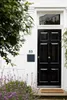 A black mailbox is mounted on a white wall next to a front door, with purple flowers in the foreground and green foliage on the left. Náhled obrázku produktu