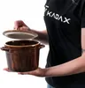 A person is holding a brown ceramic pot with a lid. Produktbild-Vorschau