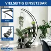 A black metal plant stand with multiple shelves, filled with various potted plants, displayed indoors against a white wall. A dining table with white tablecloth and chairs is visible in the background. Produktbild-Vorschau