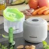A white electric mini chopper with a green lid and blades is shown on a dark surface, with various vegetables and almonds in the background. Náhľad obrázku produktu