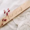 A wooden box with engraved text and decorative leaves, placed next to a baby's white dress and dried flowers. Voir les images du produit