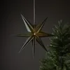 A dark green, seven-pointed star-shaped lantern hangs in front of a dark gray background, with a portion of a green Christmas tree visible on the right. Podgląd zdjęcia produktu