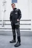 A man wearing a black and gray work overall stands in front of a white wall with metal pipes. Produktbild-Vorschau