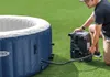 A man is setting up an inflatable hot tub outdoors on a grassy surface, connecting a pump unit to the tub. Produktbild-Vorschau