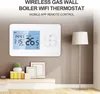 A white smart thermostat is mounted on a light brown wall, with a floor lamp and decorative items on a shelf below. Podgląd zdjęcia produktu
