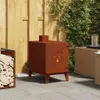 A rusty corten steel garden stove is shown on a paved patio next to a wooden fence and some greenery. A wicker table with a glass of water is also visible. Náhľad obrázku produktu