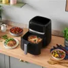 A black air fryer is shown on a wooden countertop, with food in the foreground and background. Produktbild-Vorschau