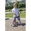 A young boy is standing on a cobblestone path holding a red balance bike. He is wearing a white polo shirt and denim shorts. The background shows a garden with green bushes and a building. Produktbild-Vorschau