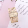 A gold-colored watch with a rectangular face and a diamond-studded bezel is placed on a pink surface next to a piece of paper with text on it. Produktbild-Vorschau