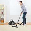 A woman is vacuuming a carpet in a well-lit room with a bookshelf in the background. Produktbild-Vorschau