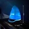 A humidifier is placed on a wooden surface next to a crib, with blue light emanating from it. Produktbild-Vorschau