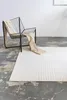 A modern chair with a beige throw draped over it sits next to a white rug with a geometric pattern on a concrete floor. Voir les images du produit