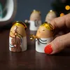 A hand is holding a small angel figurine playing a trumpet, with other angel figurines in the background. The figurines are on a wooden surface, with a blurred background suggesting a festive setting. Produktbild-Vorschau