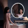 A woman applying makeup in front of a lighted, round bathroom mirror with water droplets on its surface. Produktbild-Vorschau