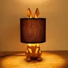 A golden rabbit-shaped table lamp with a black fabric shade sits on a wooden surface. Produktbild-Vorschau