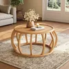 A round wooden coffee table with a curved frame base is placed on a patterned rug in a living room setting. A vase of flowers and a lit candle are on the table, with a sofa and a plant visible in the background. Anteprima dell’immagine prodotto